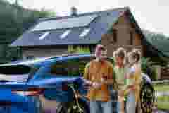 Family with little girl standing in front of their house with solar panels on the roof, having electric car.