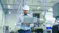 Chief Engineer in the Hard Hat Walks Through Light Modern Factory While Holding Laptop. Successful, Handsome Man in Modern Industrial Environment.
