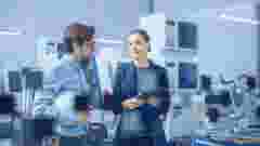 Modern Factory: Female Project Manager and Male Engineer Standing, Talking, Using Digital Tablet for Programming and Monitoring Assembly Line. Industry 4.0 Facility with High Tech Machinery